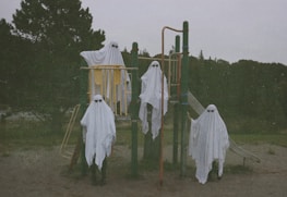 Four figures dressed in white sheets resembling ghosts stand on and around a playground structure. The setup includes a slide and climbing bars, and the background is an outdoor setting with trees and overcast skies.