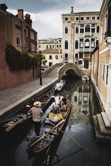 A picturesque canal with two gondolas navigating through a narrow waterway surrounded by historic buildings. The gondolier in a striped shirt maneuvers the boat with a long oar, while passengers sit and enjoy the serene atmosphere. The canal is lined with rustic buildings featuring archways, wooden shutters, and a small bridge in the background.