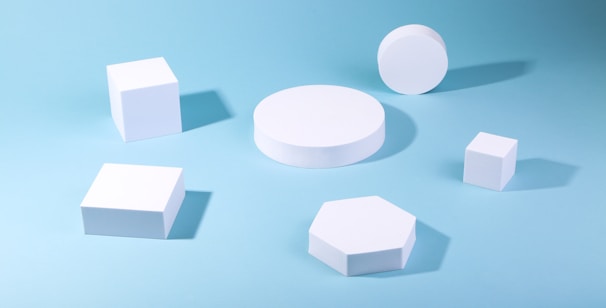 Various white geometric shapes are arranged on a light blue background, including cubes, a cylinder, a hexagon, and a circle. Shadows are cast on the surface, adding depth to the composition.