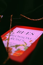 A perfume box with floral patterns and the words 'Blossom Roses' is adorned with a delicate golden chain draped over it. The box rests against a dark background, giving it a classic and elegant appearance.