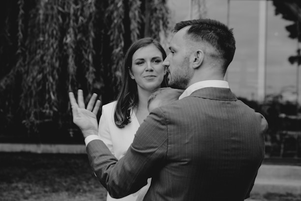 A black and white photo of a man in a suit holding a baby, with a woman standing nearby wearing a light-colored outfit. The background features trees and a reflective glass surface.