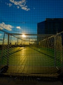 A soccer goal net is set against a vibrant sunset with a clear blue sky. The sun is partially obscured by the net and casts long shadows across the green playing field. Buildings can be seen in the background, adding an urban element to the scene.