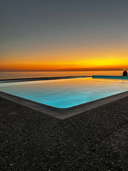 A serene swimming pool at sunset with gentle ripples reflecting red, blue, and gold hues.