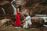 A man kneeling and proposing to a woman wearing a red dress in front of a natural waterfall setting. The scene is intimate and romantic, with lush greenery and cascading water in the background.
