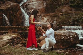 A man kneeling and proposing to a woman wearing a red dress in front of a natural waterfall setting. The scene is intimate and romantic, with lush greenery and cascading water in the background.