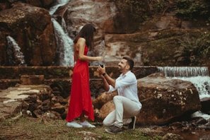 A man kneeling and proposing to a woman wearing a red dress in front of a natural waterfall setting. The scene is intimate and romantic, with lush greenery and cascading water in the background.