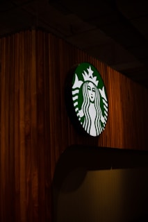 A circular sign with a green and white logo depicting a mermaid with a crown is mounted on a wooden panel wall. The lighting is dim, highlighting the texture of the wood and the logo.