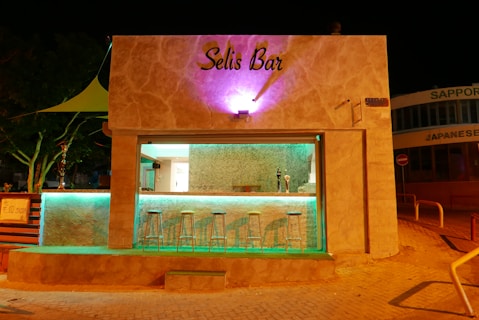 A small bar with a modern, minimalist design lit up at night. The sign reads 'Selis Bar' above an open counter area. There are several metal bar stools lined up in front of the bar, which is illuminated with turquoise lighting. The building is made of beige stone, and ambient purple lighting highlights the upper part. Trees and a canopy are partially visible to the side, while a Japanese restaurant sign is seen in the background.