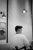 A person wearing a mask and a uniform, possibly a waiter, stands with their back turned. The setting appears to be indoors, with patterned wallpaper and industrial-style pendant lights. A framed picture hangs on the wall, and a ribbed booth is partially visible.