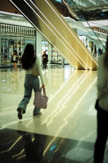 A modern shopping mall with a gleaming polished floor reflects the ambient light. Several people, including a woman holding a pink bag, are walking through the atrium. A large escalator dominates the background, next to storefronts showcasing various displays of products.