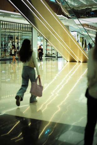 A modern shopping mall with a gleaming polished floor reflects the ambient light. Several people, including a woman holding a pink bag, are walking through the atrium. A large escalator dominates the background, next to storefronts showcasing various displays of products.