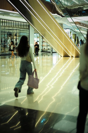 A modern shopping mall with a gleaming polished floor reflects the ambient light. Several people, including a woman holding a pink bag, are walking through the atrium. A large escalator dominates the background, next to storefronts showcasing various displays of products.