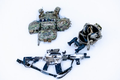 A tactical gear setup is laid out on a snow-covered surface, including a camouflage ballistic vest with pouches, a helmet with mounted night-vision goggles, and a camouflaged rifle partially covered in snow.