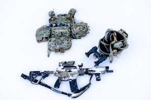 A tactical gear setup is laid out on a snow-covered surface, including a camouflage ballistic vest with pouches, a helmet with mounted night-vision goggles, and a camouflaged rifle partially covered in snow.