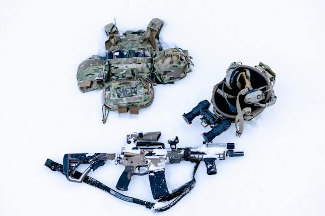 A tactical gear setup is laid out on a snow-covered surface, including a camouflage ballistic vest with pouches, a helmet with mounted night-vision goggles, and a camouflaged rifle partially covered in snow.