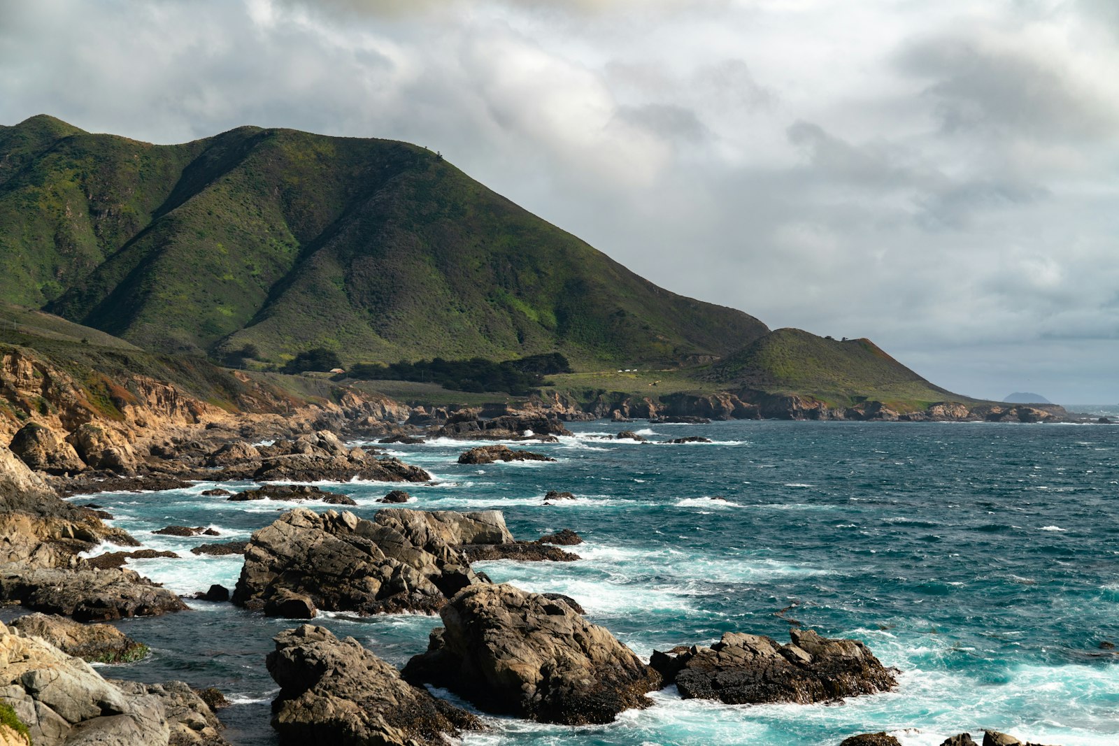 Rocky Big Sur coastline with crashing waves and green hills, brooding sky