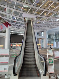 An escalator ascends towards a ceiling with a grid-like glass and metal structure. There are signs and informational panels on either side of the escalator. The setting appears to be a modern, spacious indoor public area, possibly an airport or large transportation hub.