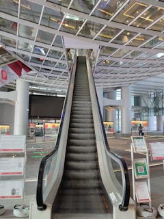 An escalator ascends towards a ceiling with a grid-like glass and metal structure. There are signs and informational panels on either side of the escalator. The setting appears to be a modern, spacious indoor public area, possibly an airport or large transportation hub.