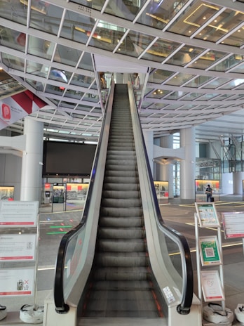 An escalator ascends towards a ceiling with a grid-like glass and metal structure. There are signs and informational panels on either side of the escalator. The setting appears to be a modern, spacious indoor public area, possibly an airport or large transportation hub.