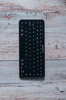 A black Bluetooth keyboard with circular keys is placed on a rustic wooden surface. The keyboard includes standard keys such as QWERTY layout, numbers, and function keys. The texture of the wooden surface features a weathered, worn look with variations in shades of light brown and white.