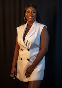 A woman is smiling broadly, wearing a stylish white sleeveless blazer dress with black buttons. She has long dark hair and is accessorized with hoop earrings and rings on her fingers. The background is dark, adding contrast to her outfit.