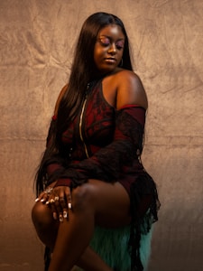 A woman with long straight hair sits gracefully on a textured surface. She wears a black and red lace outfit with intricate patterns. Her eyes are closed, showing purple eyeshadow, and her pose exudes confidence. Her nails are long and manicured, adorned with rings and designs.