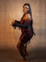 A woman stands against a plain, textured backdrop looking at the camera with a big smile. She is wearing a black and red off-shoulder dress with lace details and has long, straight black hair. Her pose is dynamic, with one leg slightly raised and her hands gesturing as if she is mid-dance or showing off her nails.