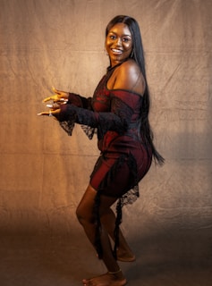 A woman stands against a plain, textured backdrop looking at the camera with a big smile. She is wearing a black and red off-shoulder dress with lace details and has long, straight black hair. Her pose is dynamic, with one leg slightly raised and her hands gesturing as if she is mid-dance or showing off her nails.