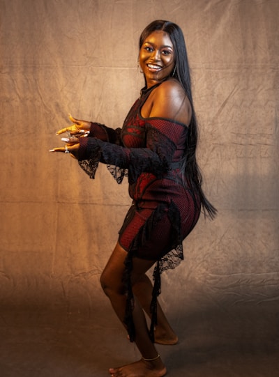 A woman stands against a plain, textured backdrop looking at the camera with a big smile. She is wearing a black and red off-shoulder dress with lace details and has long, straight black hair. Her pose is dynamic, with one leg slightly raised and her hands gesturing as if she is mid-dance or showing off her nails.