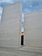 Two massive, vertical concrete slabs stand parallel to each other with a gap between them, through which part of a yellow building with a red roof is visible. The sky overhead is partly cloudy, creating a backdrop of blue and gray hues. The ground and the slabs are made of the same textured material.