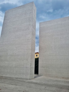 Two massive, vertical concrete slabs stand parallel to each other with a gap between them, through which part of a yellow building with a red roof is visible. The sky overhead is partly cloudy, creating a backdrop of blue and gray hues. The ground and the slabs are made of the same textured material.