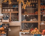 A rustic market stall displaying a variety of fresh produce and goods. Grapes, bell peppers, citrus fruits, and assorted vegetables are neatly arranged in wooden crates and cardboard boxes. Jars and bottles are visible on the shelves behind the produce. The stall is framed by an overhanging branch with yellowing leaves, adding an autumnal feel.