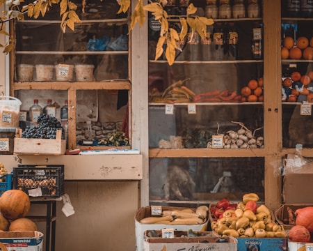 A rustic market stall displaying a variety of fresh produce and goods. Grapes, bell peppers, citrus fruits, and assorted vegetables are neatly arranged in wooden crates and cardboard boxes. Jars and bottles are visible on the shelves behind the produce. The stall is framed by an overhanging branch with yellowing leaves, adding an autumnal feel.