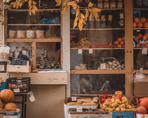 A rustic market stall displaying a variety of fresh produce and goods. Grapes, bell peppers, citrus fruits, and assorted vegetables are neatly arranged in wooden crates and cardboard boxes. Jars and bottles are visible on the shelves behind the produce. The stall is framed by an overhanging branch with yellowing leaves, adding an autumnal feel.