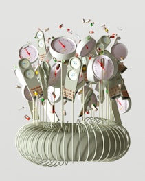 A collection of devices resembling clocks or gauges with colored indicators is attached to long, thin rods emerging from a coil structure. The devices have dials with red needles and various numerical scales. Surrounding the devices are individual pills and capsules floating through the air, giving a sense of motion.