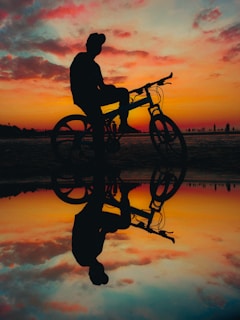 A sunset silhouette of a rider taking a break, sitting on a log with their bike.