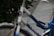 A close-up of a bicycle frame with a gradient from white to blue, displaying the brand name 'DIANA'. The bicycle features a metallic wheel rim and a sticker with a winged logo. Green leaves and pavement are visible in the background.