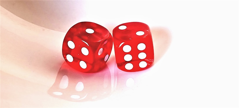 Two red translucent dice sit side by side on a reflective surface. Each die displays a different combination of white dots, representing numbers. The background is a clean, light color that enhances the brightness of the red hues.
