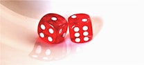 Two red translucent dice sit side by side on a reflective surface. Each die displays a different combination of white dots, representing numbers. The background is a clean, light color that enhances the brightness of the red hues.