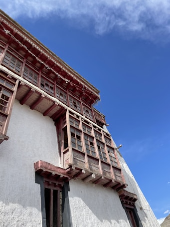 A traditional building facade with multiple wooden-framed windows and an intricate wooden overhang, set against a bright blue sky. The white plastered wall features decorative textural patterns, enhancing the architectural detail.