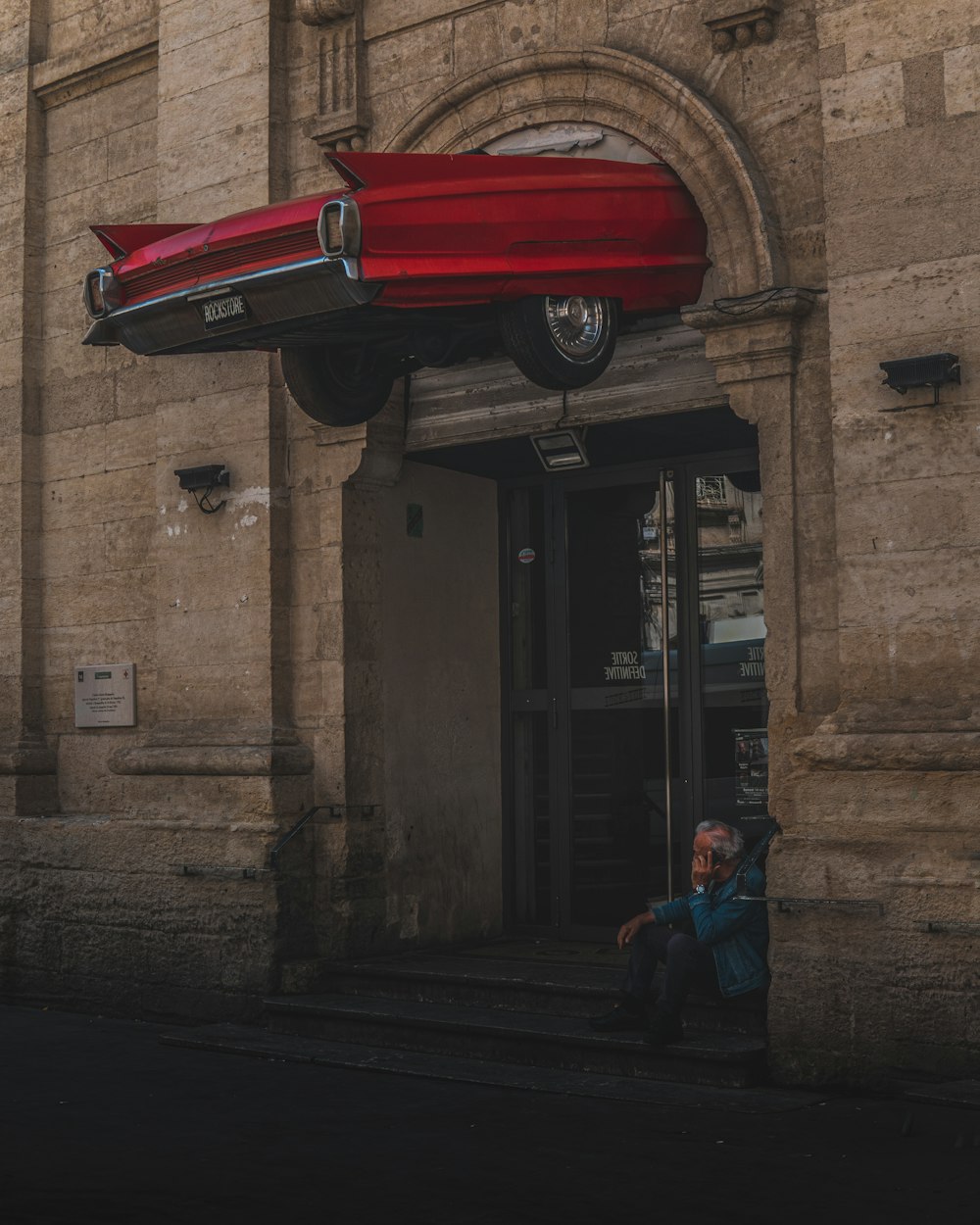 a person sitting on the steps of a building with a red car flying over him