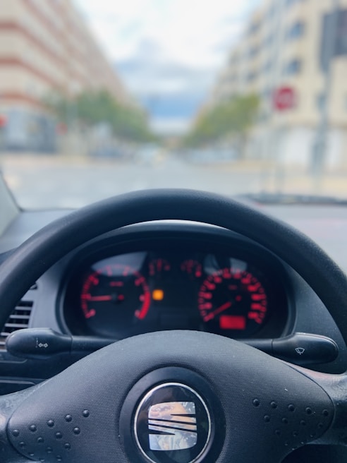 A car dashboard is visible from the driver's perspective, with the steering wheel prominently in the foreground. The dashboard displays various gauges and indicators, including speedometer, tachometer, and fuel gauge, with red and white markings. The background is blurred, showing an out-of-focus urban street scene with buildings on either side.