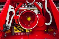 A close-up view of a complex mechanical system primarily composed of red metal structures. The center features a cylindrical component with a bright yellow detail inside. The surrounding framework includes various geometric shapes and details, with some parts painted in white. Cables and chains are visible, adding to the industrial feel.