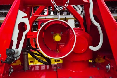 A close-up view of a complex mechanical system primarily composed of red metal structures. The center features a cylindrical component with a bright yellow detail inside. The surrounding framework includes various geometric shapes and details, with some parts painted in white. Cables and chains are visible, adding to the industrial feel.