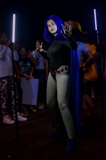 A person dressed in a detailed and elaborate cosplay outfit stands in front of two vertical LED lights, engaging with the camera. The person is wearing a blue wig, a blue cape, and a costume with red gems. Other people in casual clothing are milling around in the background, some observing the cosplayer and possibly holding posters or papers.