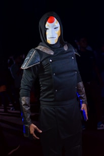 A person dressed in a dark, armored outfit with metallic shoulder pads and forearm guards, wearing a distinctive white mask with red accents. The ensemble is complemented by a hood, giving an overall mysterious and enigmatic appearance, set against a dimly lit background with other figures partially visible.