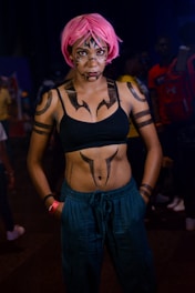 A person with bold, artistic body paint and a pink wig stands confidently against a dark background. The paint includes intricate designs on the face, chest, and arms. They wear a black crop top and blue pants, exuding a sense of style and creativity. Other people are visible in the background, adding a subtle sense of a social environment.