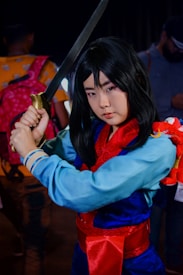 A person cosplaying as a warrior character, holding a sword with both hands. They are wearing a traditional outfit with a blue tunic, red sash, and patterned red scarf. A small red dragon toy is attached to their arm. The background features other people and appears to be a crowded indoor event.