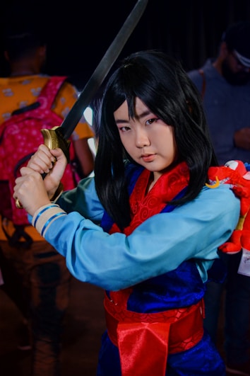 A person cosplaying as a warrior character, holding a sword with both hands. They are wearing a traditional outfit with a blue tunic, red sash, and patterned red scarf. A small red dragon toy is attached to their arm. The background features other people and appears to be a crowded indoor event.