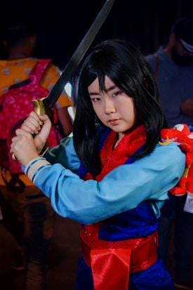 A person cosplaying as a warrior character, holding a sword with both hands. They are wearing a traditional outfit with a blue tunic, red sash, and patterned red scarf. A small red dragon toy is attached to their arm. The background features other people and appears to be a crowded indoor event.